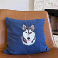 Load image into Gallery viewer, A square blue throw pillow featuring a detailed custom embroidered husky dog face, resting on a brown leather armchair in front of a stone fireplace.
