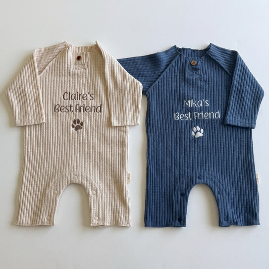 Two ribbed long-sleeve baby footless onesies laid flat on a light background. The left one is beige with the text 'Claire's Best friend' and a dark paw print, and the right one is navy blue with the text 'Mika's Best friend' and a white paw print.
