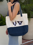 Load image into Gallery viewer, A woman wearing a navy sleeveless top and light blue jeans carries a large cream canvas tote bag with a navy bottom panel and printed Boston Terrier dog faces on the front, set against an outdoor patio background with potted plants.
