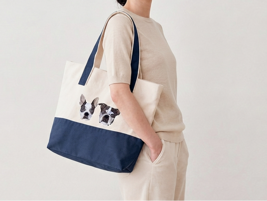 A woman wearing a beige outfit carries a canvas tote bag with navy blue straps and bottom panel. The front of the bag is cream with custom embroidered dog portrait of two Boston Terriers.