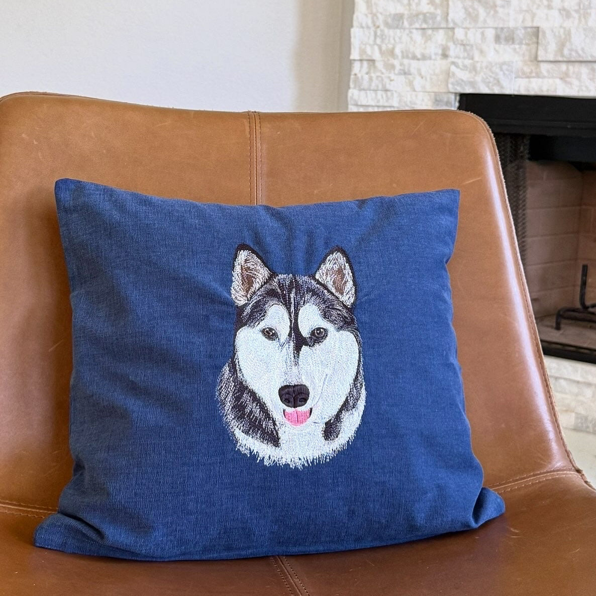 A square blue throw pillow featuring a detailed custom embroidered husky dog face, resting on a brown leather armchair in front of a stone fireplace.